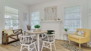 Una habitación luminosa y bien iluminada con una mesa redonda para comer, sillas blancas, un sillón amarillo y una chimenea.