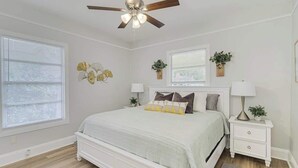 Un dormitorio con cama, mesita de noche, ventilador de techo, ventana con persianas y una planta en la pared.