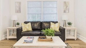 Una sala de estar con un sofá de cuero negro, dos mesitas de noche blancas con lámparas, una mesa de centro con revistas y una ventana con persianas.