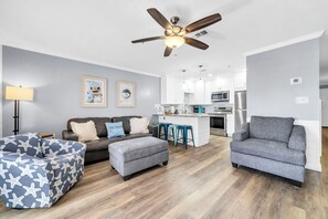 A modern living room with a sofa, ottoman, and armchair, a ceiling fan, and a kitchen area with white cabinets and appliances.