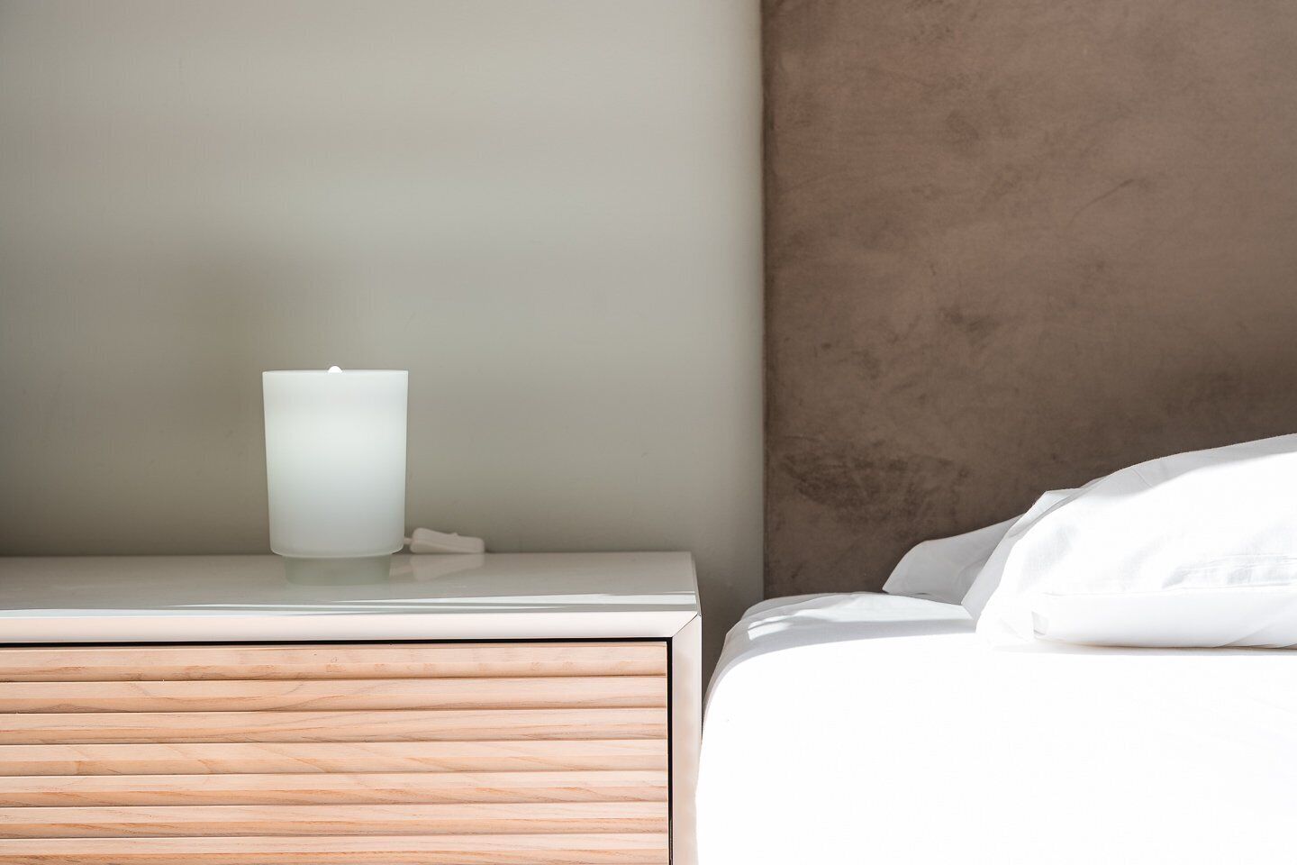 Een minimalistische slaapkamer met een houten nachtkastje, een witte bijzettafel en een bed met witte lakens.