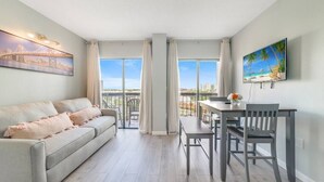 Ein modernes Wohnzimmer mit Sofa, Esstisch und Stühlen, großen Fenstern mit Vorhängen und Kunstwerken an den Wänden.