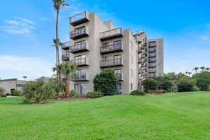 Um edifício residencial com vários andares e varandas, rodeado por um relvado bem cuidado e palmeiras.