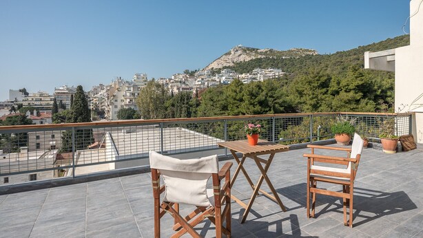 Una terraza en la azotea con dos sillas de madera y una mesa, con vista a un paisaje urbano con edificios y una colina.