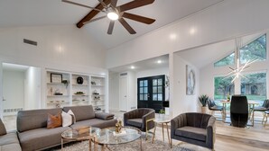 A spacious living room with a ceiling fan, a sofa, armchairs, a glass coffee table, and a bookshelf. - Lux Dallas Oasis- Pool, Hot Tub, Firepit, Game Room, Sleeps 12 (Dallas)