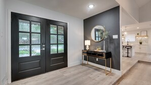 A modern entryway with a black double door, a console table with a lamp, and a round mirror on the wall. - Lux Dallas Oasis- Pool, Hot Tub, Firepit, Game Room, Sleeps 12 (Dallas)