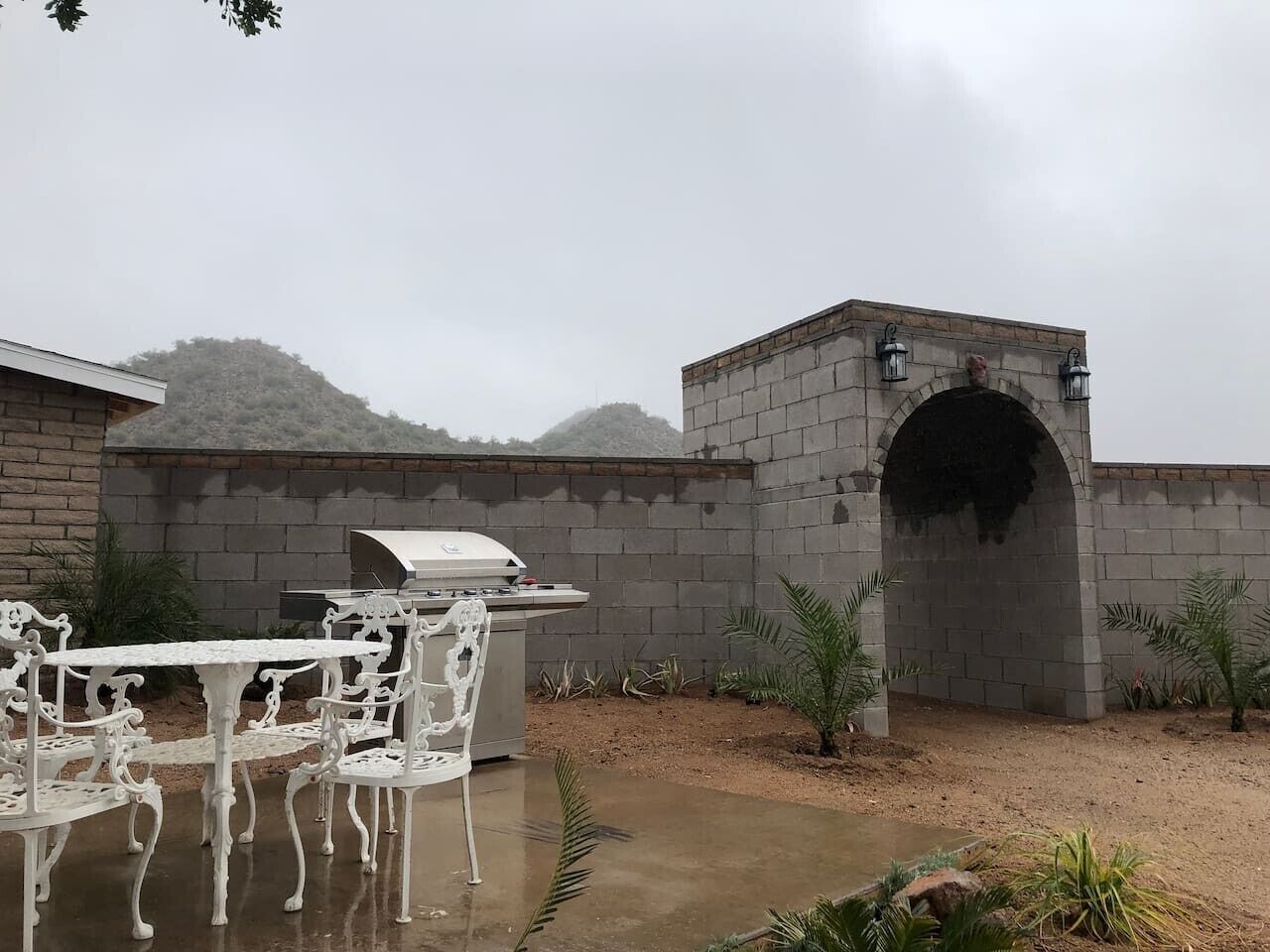 An outdoor patio with a barbecue grill, white ornate chairs, and a stone archway.