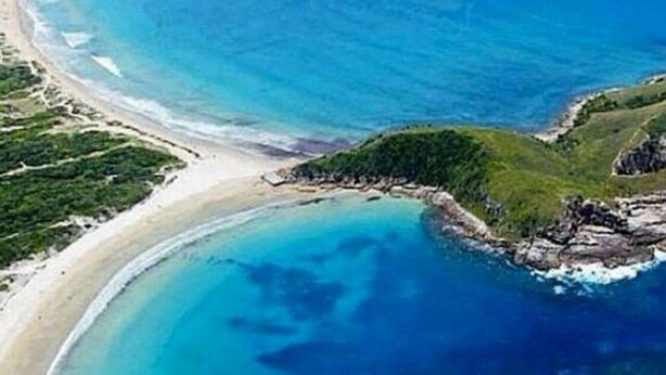 An aerial view of a coastal area with a curved bay, sandy beach, and green vegetation.