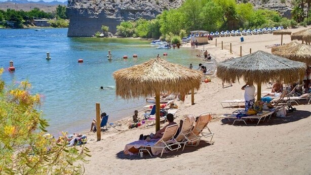 Une plage bordée de parasols en chaume, de chaises longues et dont l’arrière-plan est constitué de rochers.