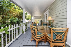 Relax on the covered porch with a glass-top table and wicker chairs, surrounded by a lush garden view.