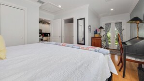 A bedroom with a large bed, a chair, a desk, and a view of the kitchen. - Downtown Dunedin Cozy 1 bedroom Cottage (Dunedin)