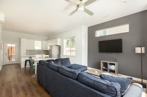 A modern living room with a sectional sofa, a dining area with chairs, a kitchen with white cabinets, a flat-screen TV mounted on the wall, and a ceiling fan.