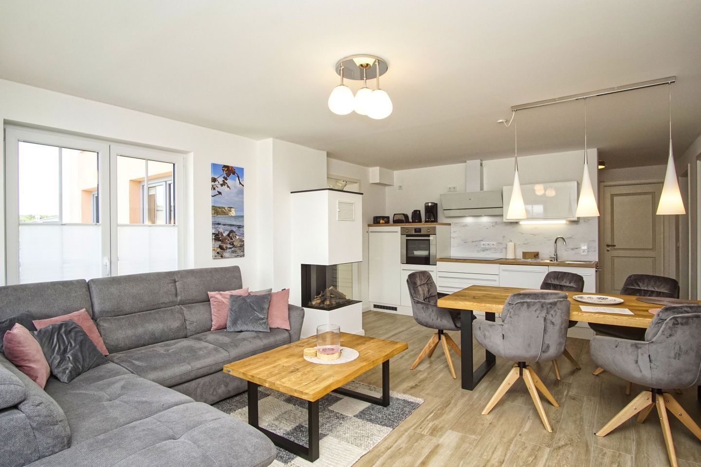 A modern living room with a grey sofa, wooden coffee table, and grey chairs. A kitchen with white cabinets and a dining table with chairs is visible in the background.