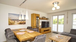 Ein modernes Wohnzimmer mit Esstisch aus Holz, grauen Polsterstühlen, einem grauen Sofa und einem Fernseher auf einer Holzkommode.