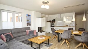 Ein modernes Wohnzimmer mit einem grauen Sofa, einem Holztisch und grauen Sesseln. Im Hintergrund ist eine Küche mit weißen Schränken und einem Esstisch mit Stühlen zu sehen.