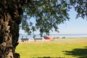 Eine Rasenfläche mit Bank und Baum, ein Gewässer und eine entfernte Ansicht eines Strandes mit Zelten.