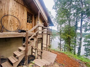 A wooden cabin with a wooden staircase leading to a deck overlooking a lake, surrounded by trees. - Pets/Kids love the Jacuzzi @ Large Lakeside Cabin (Talladega)