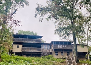 A two-story building with a balcony on the upper floor, surrounded by trees and vegetation.