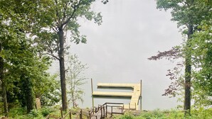 Een houten brug leidt naar een groot, modern gebouw met een plat dak, omgeven door weelderige begroeiing en bomen.