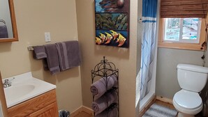Une salle de bain avec un lavabo blanc, un meuble en bois, un porte-serviettes avec des serviettes, un tableau représentant des poissons, une fenêtre avec des stores et des toilettes.