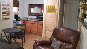 Un espace de vie compact comprenant un coin cuisine, un coin repas avec une table ronde et des chaises, et un canapé en cuir agrémenté d’un coussin à motif léopard.
