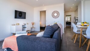 Un salon moderne avec un canapé gris, un téléviseur à écran plat fixé au mur, un coin repas avec une table et des chaises, et une cuisine en arrière-plan.