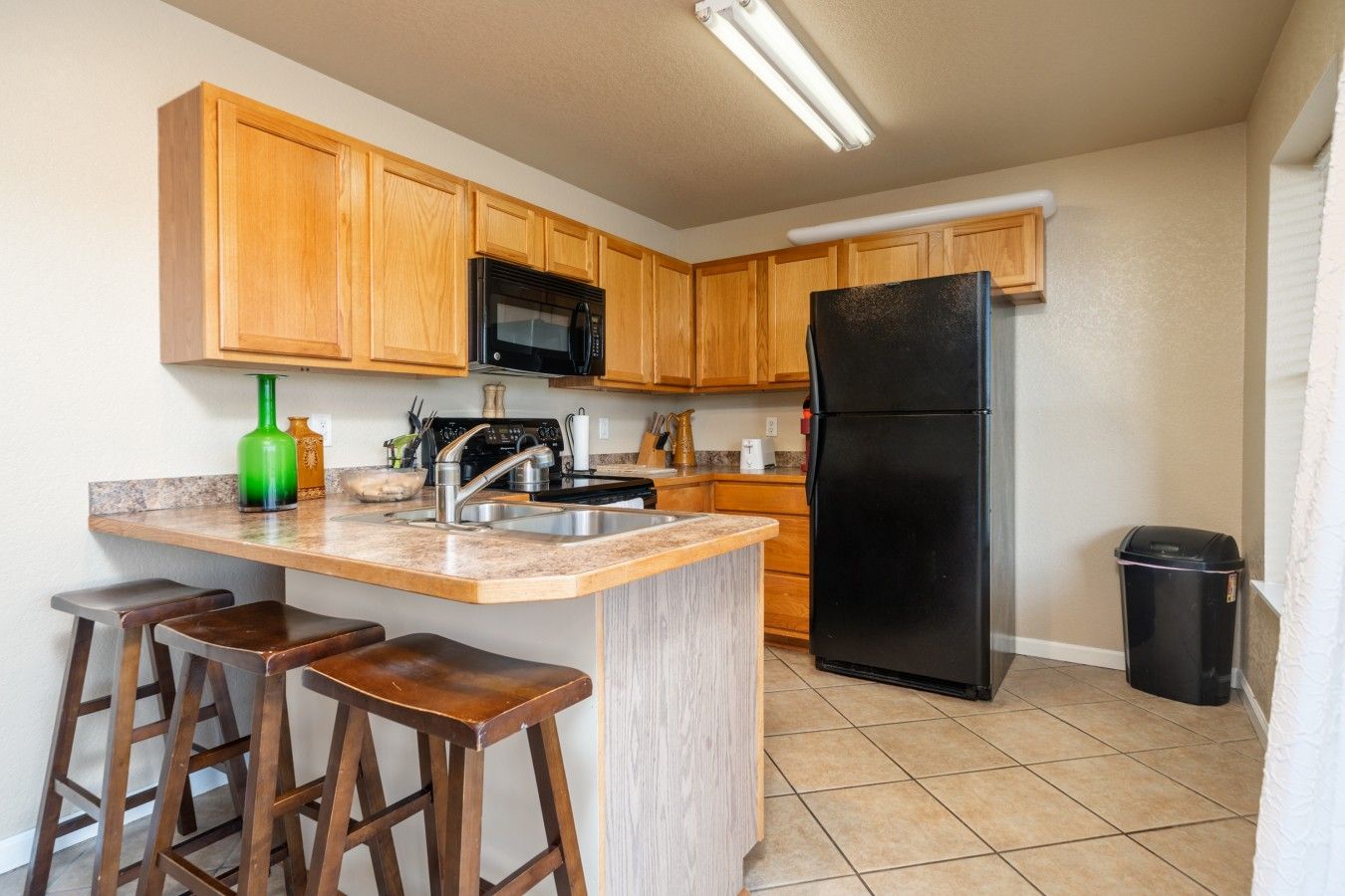A kitchen with wooden cabinets, a black refrigerator, a microwave, a sink, and a bar stool area.