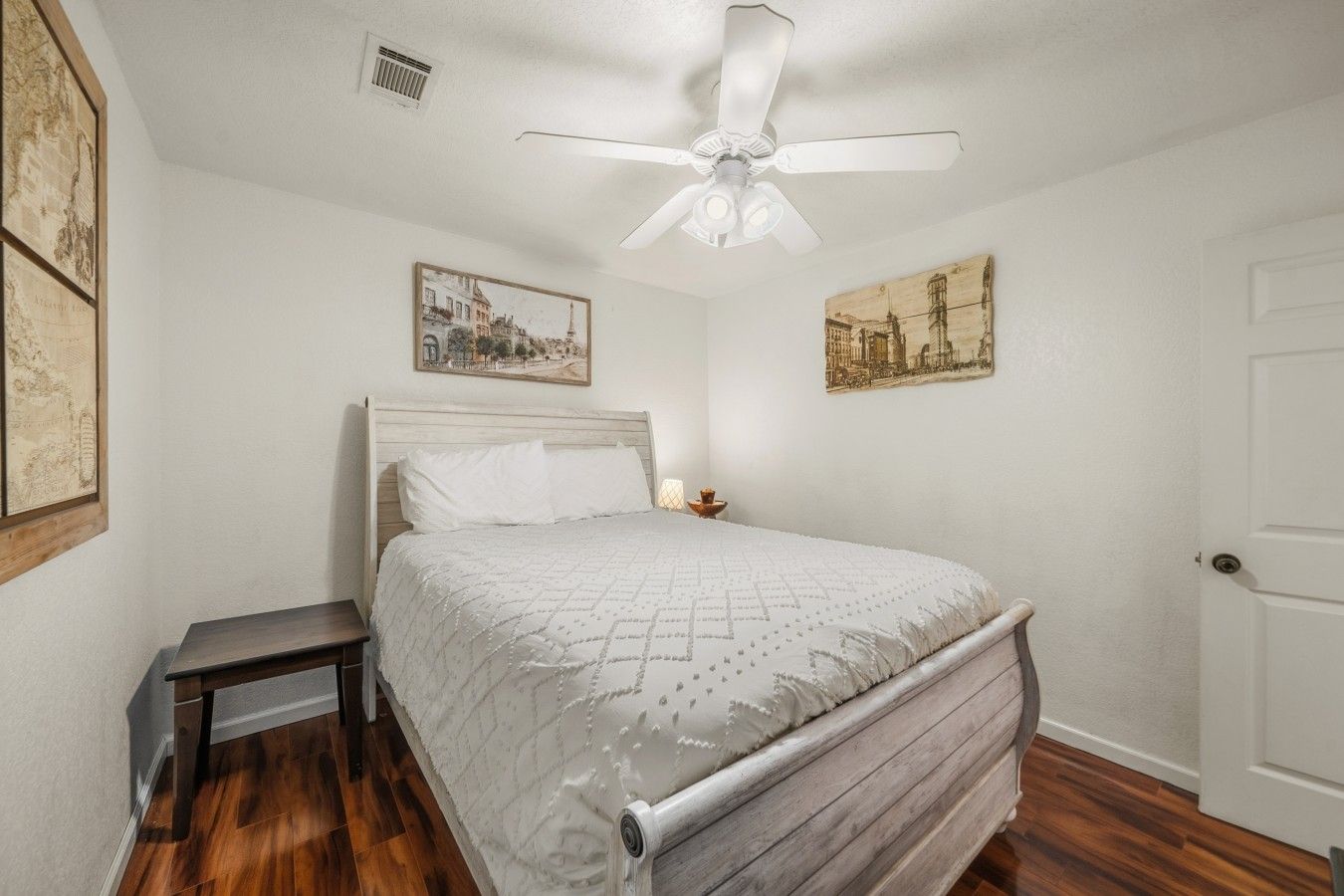 Un dormitorio con cama, mesita de noche, ventilador de techo y dos cuadros en la pared.