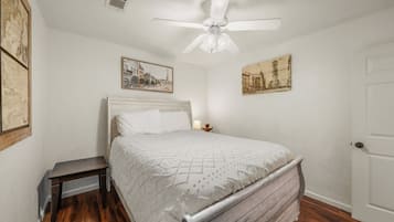 Un dormitorio con cama, mesita de noche, ventilador de techo y dos cuadros en la pared.