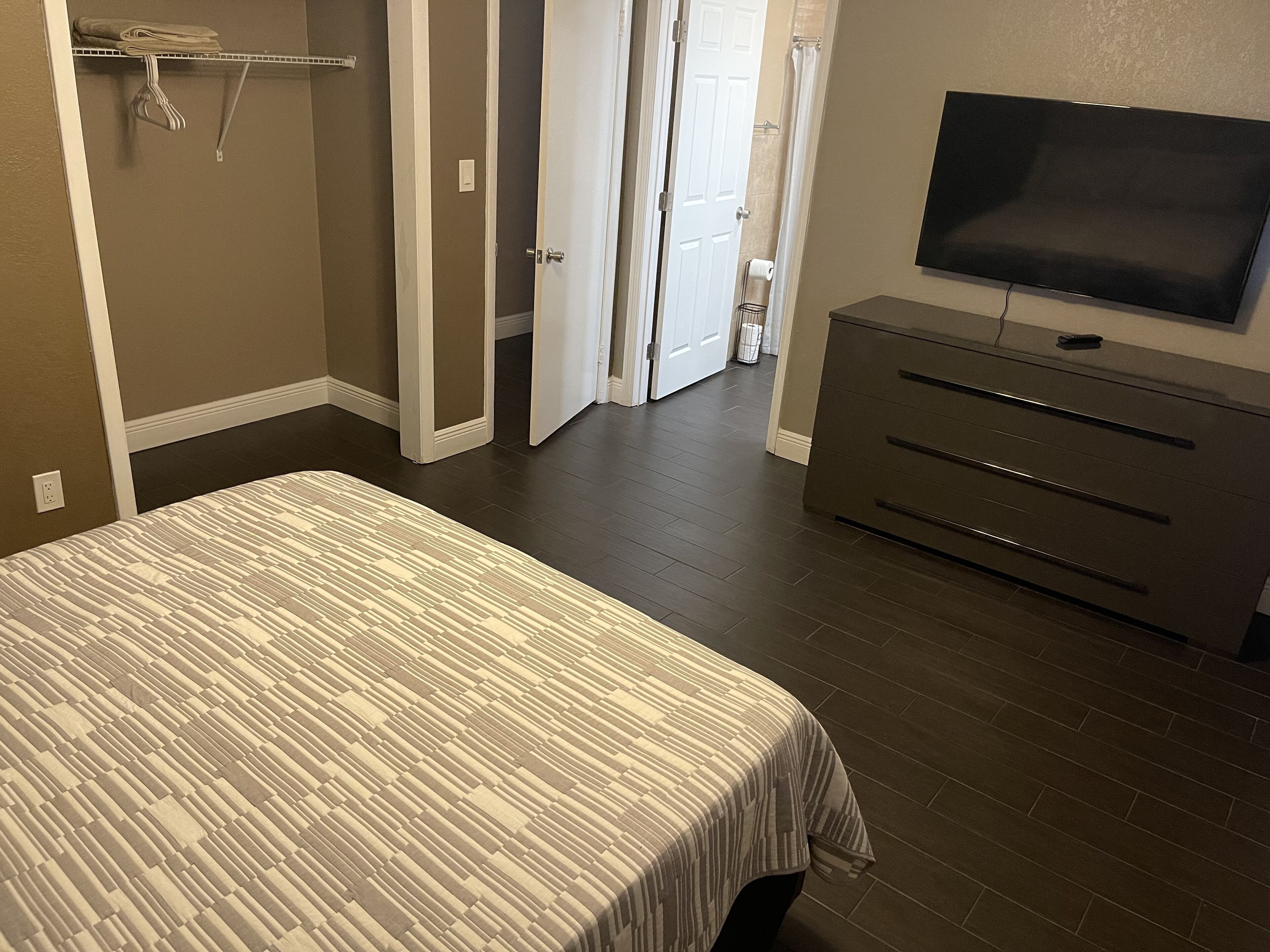 A bedroom with a bed, a dresser, a TV, and a door leading to a bathroom.