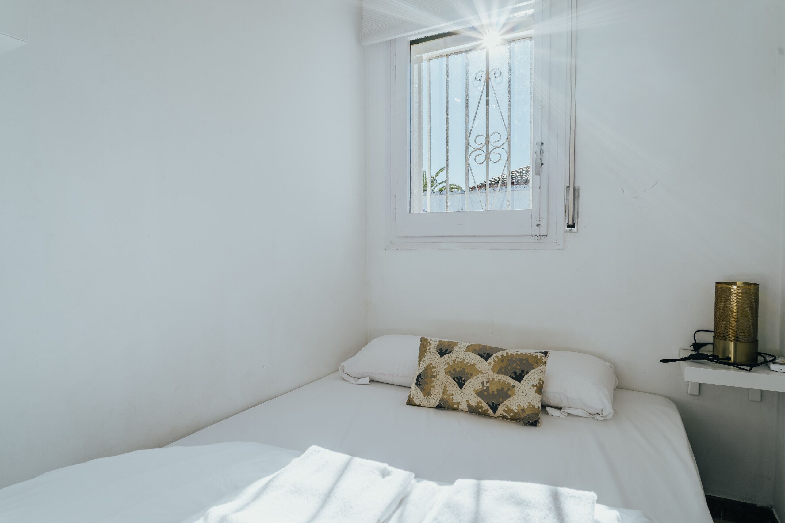 A bedroom with a bed, a window with bars, a bedside table with a lamp, and a patterned pillow.