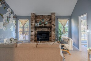 Cozy living room with a brick fireplace, comfortable sofa, and wooden chair, offering a view of the surrounding trees.