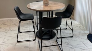 A dining area with a round table, four black chairs, a chandelier, and a window with blinds.