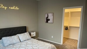 A bedroom with a large bed, a bedside table, a nightstand, a wall-mounted artwork, and a closet with a door open.