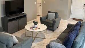A living room with a flat-screen TV on a cabinet, a round coffee table with magazines, and two grey sofas with cushions.
