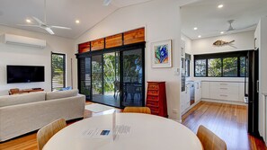A modern living space with a flat-screen TV, a dining area with a table and chairs, a kitchen with white cabinets, and a large sliding glass door leading to a balcony. - Frangipani Place - 3 BR - Beachfront (Wongaling Beach)