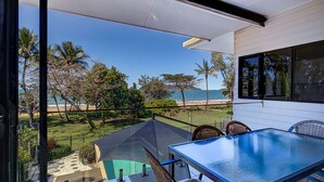 A balcony with a glass table and chairs overlooking a beach with palm trees and ocean.