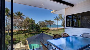 A balcony with a glass table and chairs overlooking a beach with palm trees and ocean.