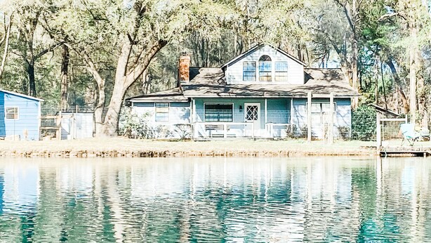Een tweelaags huis met een veranda, omringd door bomen en water.