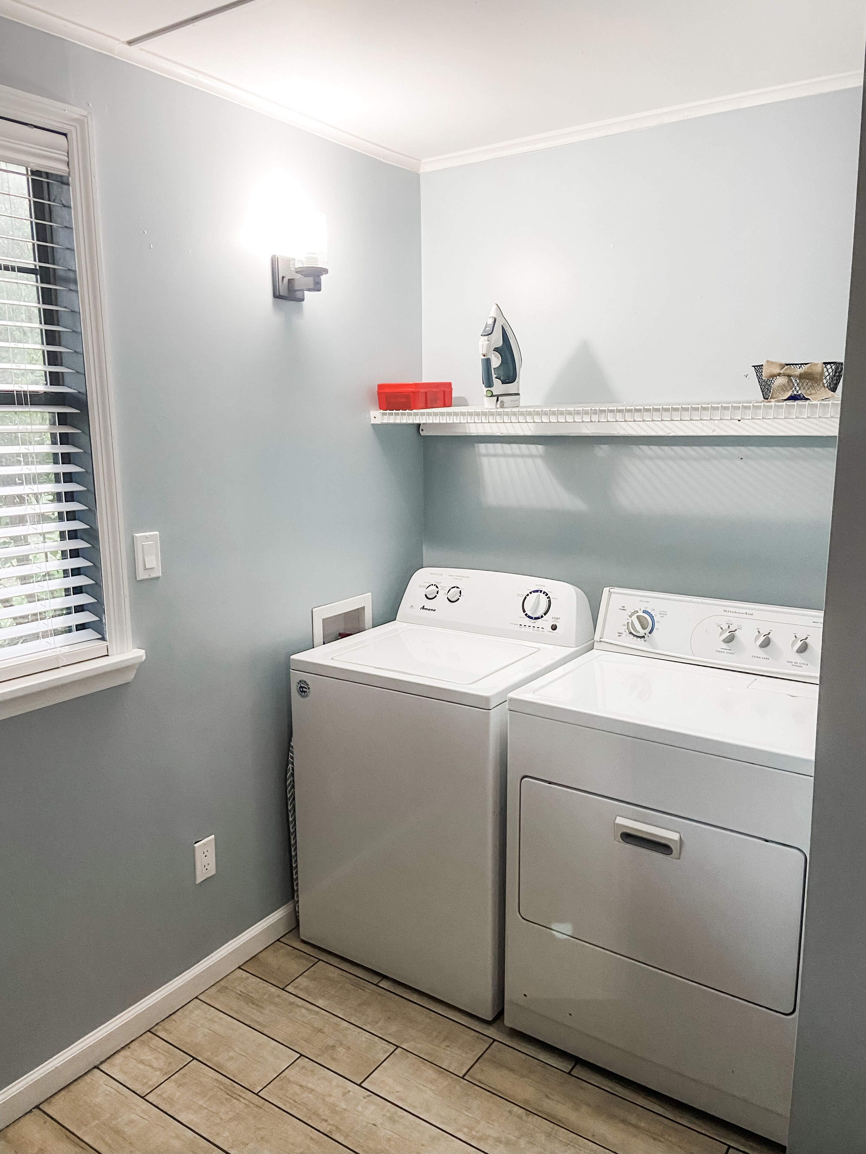 A laundry room with two white washers and dryers, a window with blinds, and a shelf with a basket and a red container.