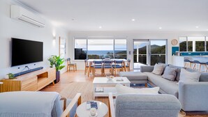 A modern living room with a flat-screen TV, a dining area with blue chairs, and a sofa set. - Becker Bliss - 5 bedrooms, ocean views, sleeps 16 (Forster)