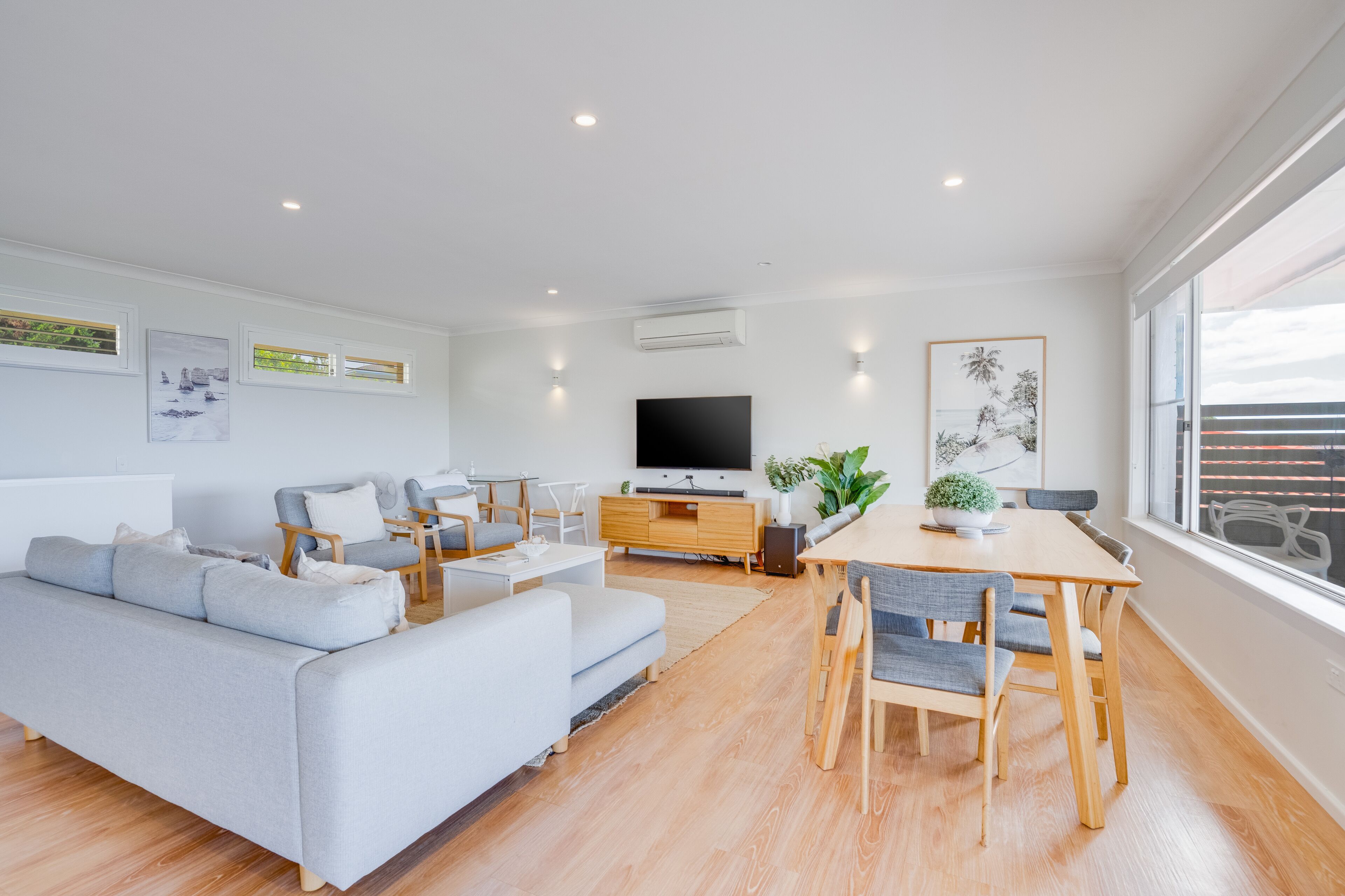 A modern living room with a grey sofa, a dining table with chairs, a flat-screen TV, and artwork on the walls.