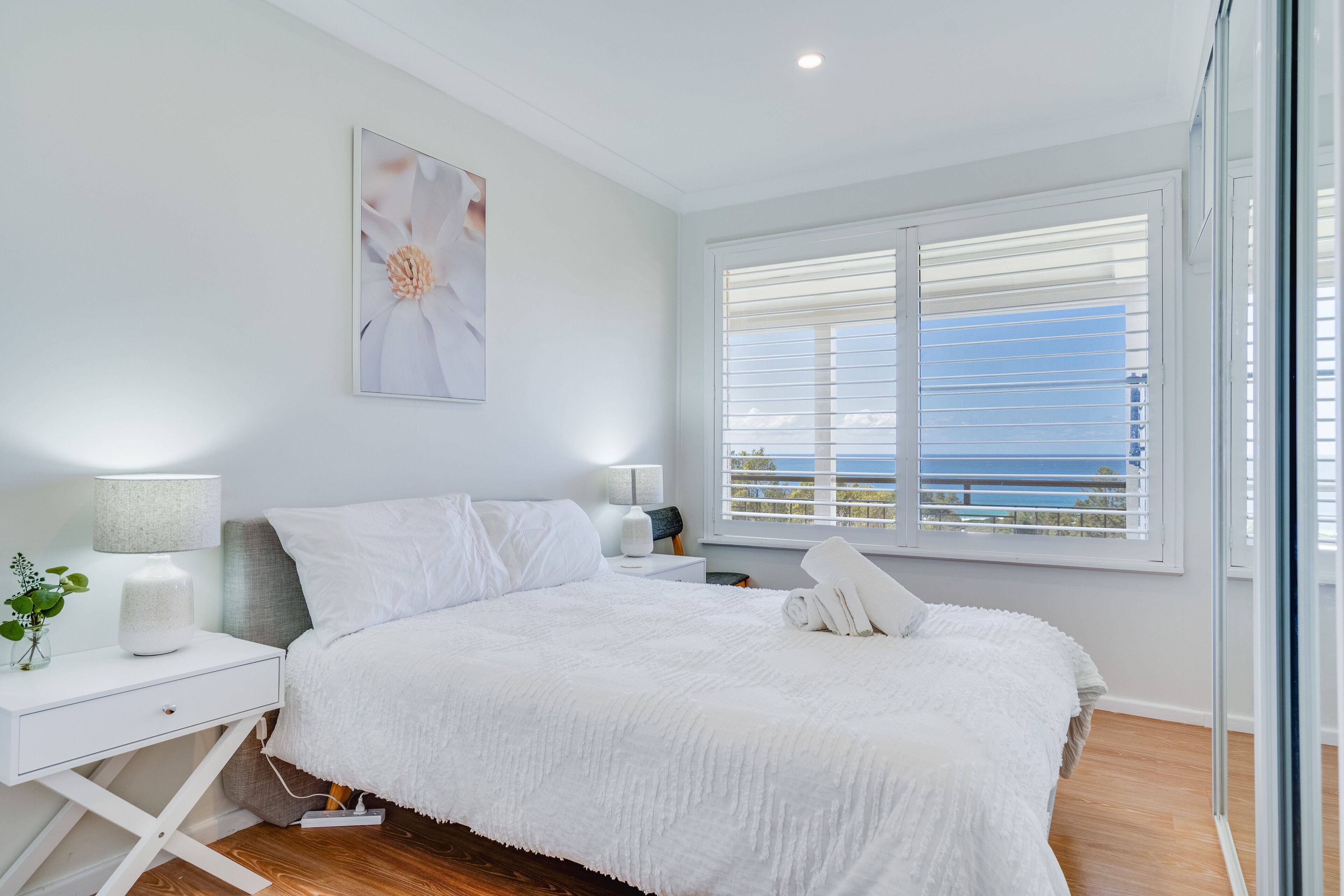 Une chambre avec un grand lit, du linge de lit blanc, une table de chevet avec une lampe, une fenêtre avec des stores et une vue sur la mer.