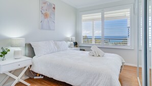 A bedroom with a large bed, white bedding, a bedside table with a lamp, a window with blinds, and a view of the sea. - Becker Bliss - 5 bedrooms, ocean views, sleeps 16 (Forster)