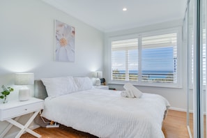 A bedroom with a large bed, white bedding, a bedside table with a lamp, a window with blinds, and a view of the sea. - Becker Bliss - 5 bedrooms, ocean views, sleeps 16 (Forster)
