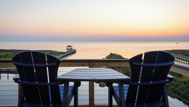 A deck with two Adirondack chairs and a table, overlooking a calm body of water and a pier. - Waterfront Home with Panoramic Views in Black Dog Harbor, Salvo, NC  (Salvo)