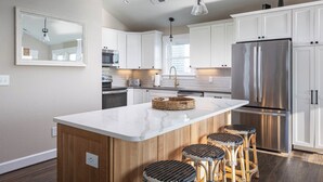 A modern kitchen with white cabinets, a stainless steel refrigerator, a marble island with bar stools, and pendant lighting. - Waterfront Home with Panoramic Views in Black Dog Harbor, Salvo, NC  (Salvo)