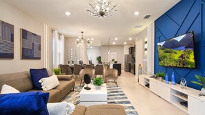 A modern living room with a sofa, coffee table, and TV. There is a dining area with chairs and a kitchen in the background. - Disney Getaway w Moana Themed Rm & More, Games! (Davenport)