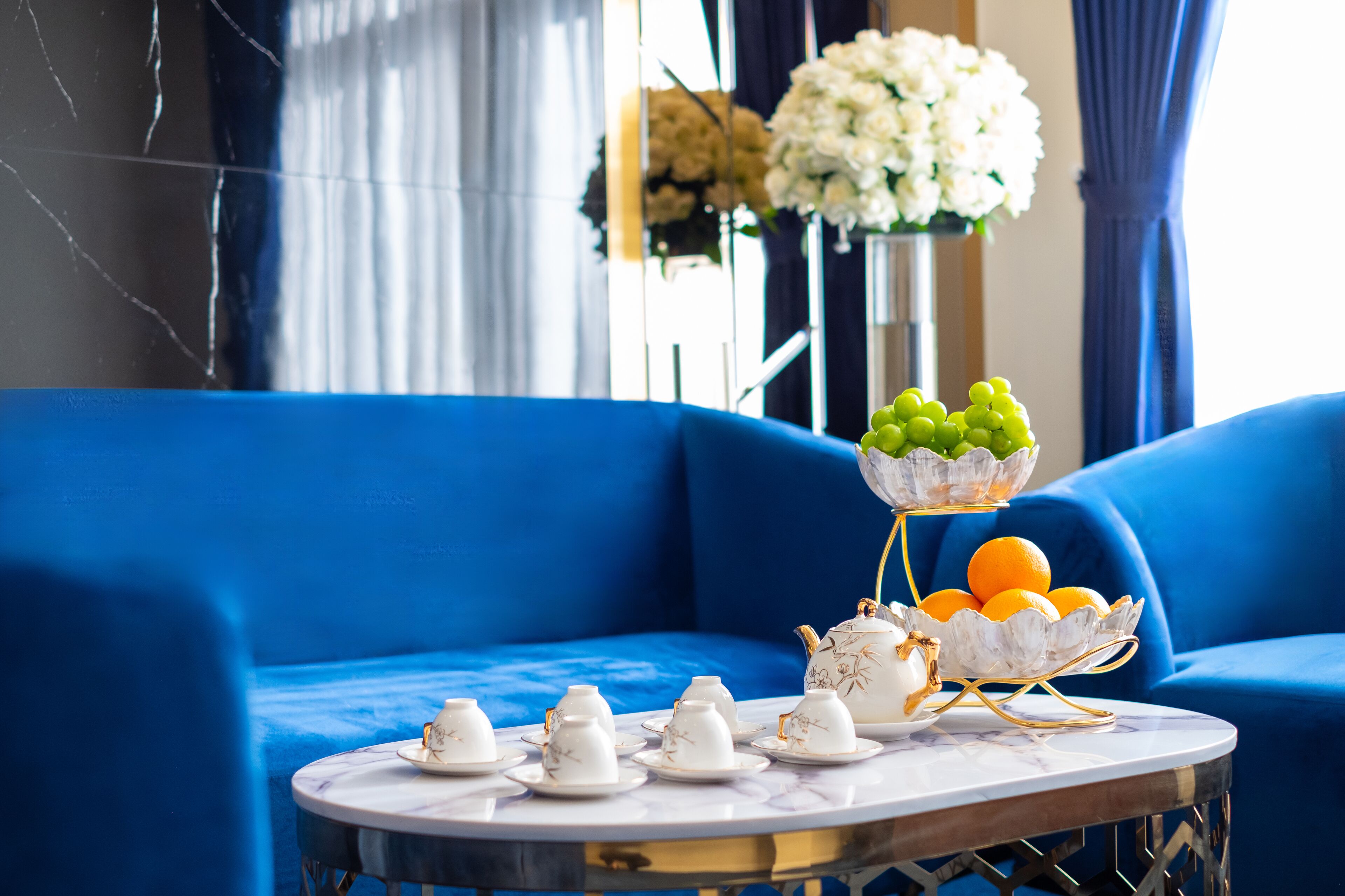 Un canapé bleu, une table ronde avec un service à thé et un saladier rempli d’oranges et de raisins.
