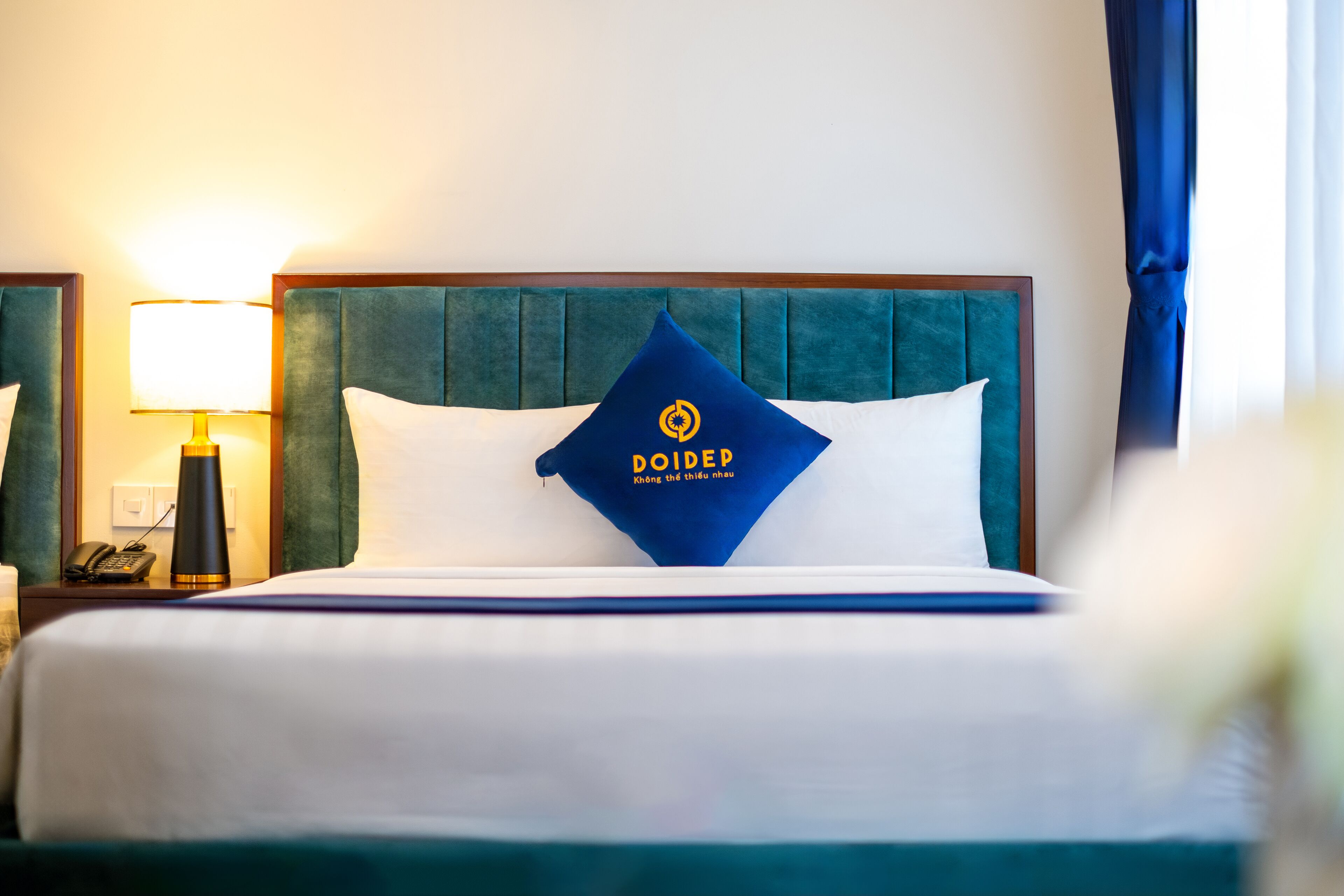 Una habitación de hotel con cama, mesita de noche con lámpara y almohada azul con texto.